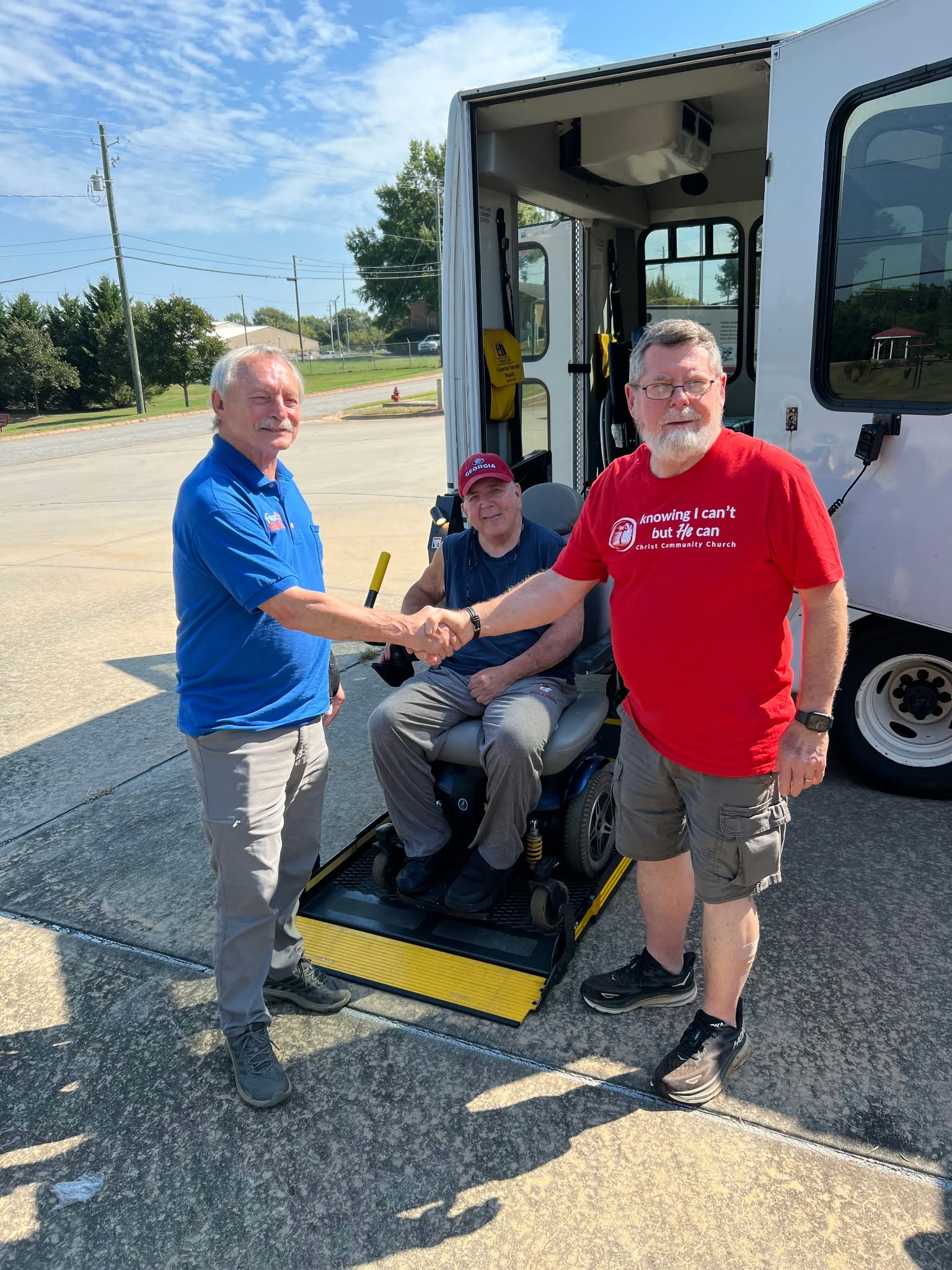 Two men shaking hands in front of an accessible van while a neighbor in a power wheelchair rides the lift ramp