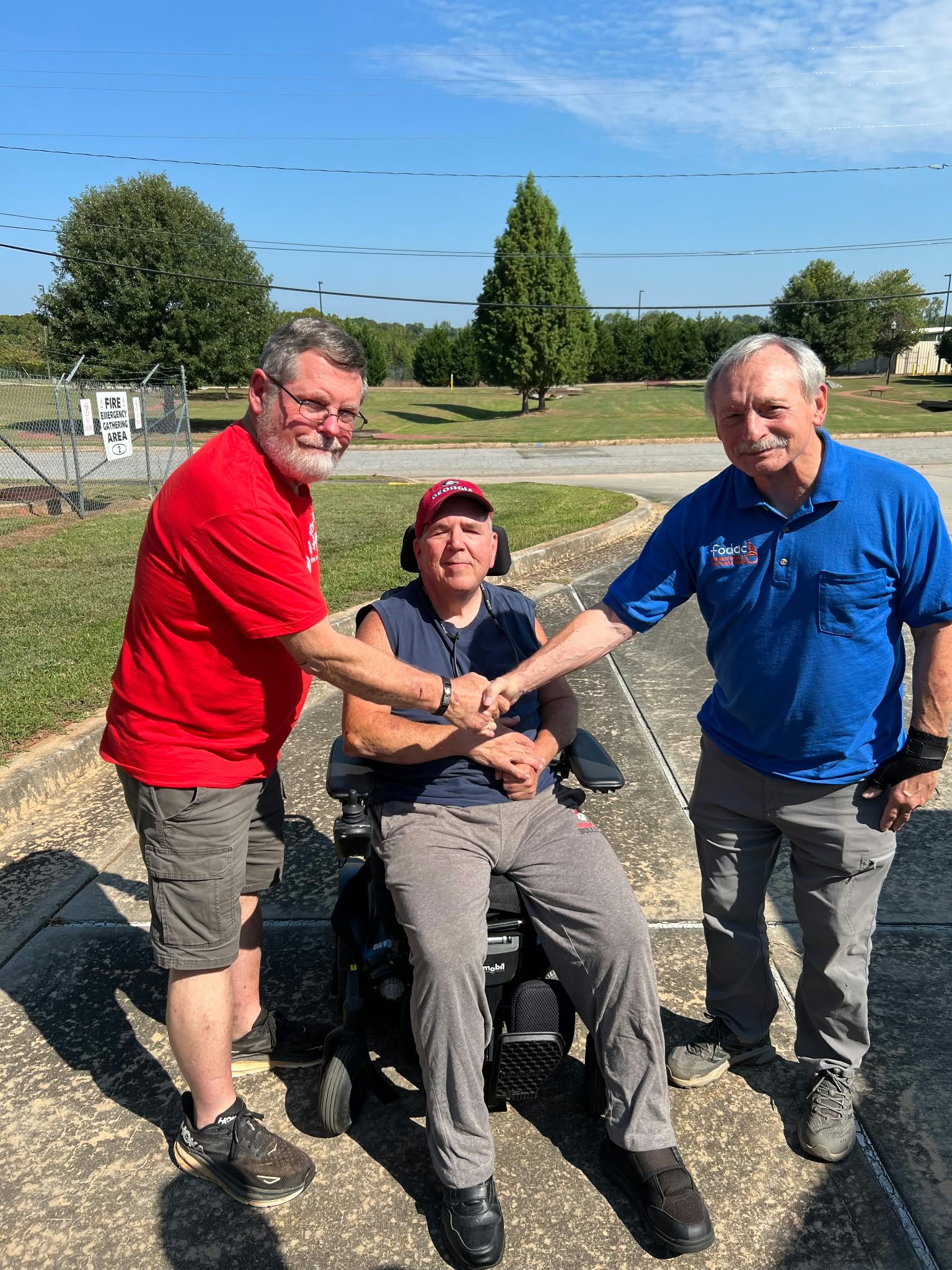 Two men shaking hands outdoors with a smiling neighbor in a power wheelchair between them